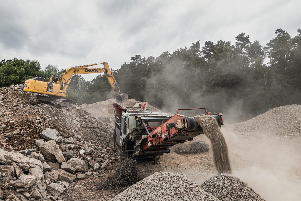 Kettenbagger belädt eine mobile Brechanlage mit Bauschutt, aus der aufbereiteter Schotter über ein Förderband austritt