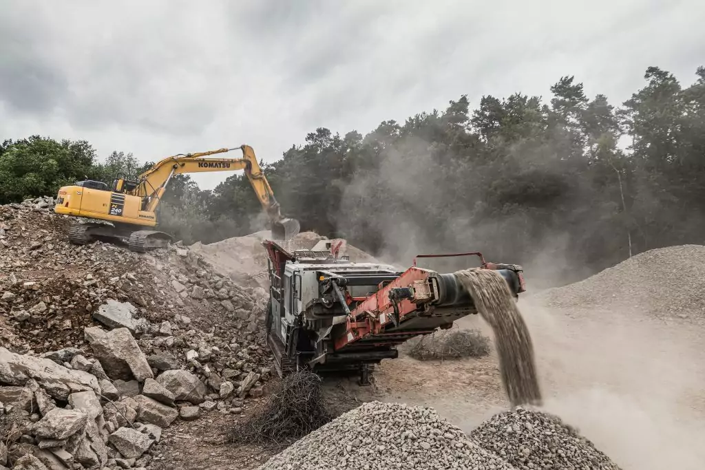 Kettenbagger belädt eine mobile Brechanlage mit Bauschutt, aus der aufbereiteter Schotter über ein Förderband austritt